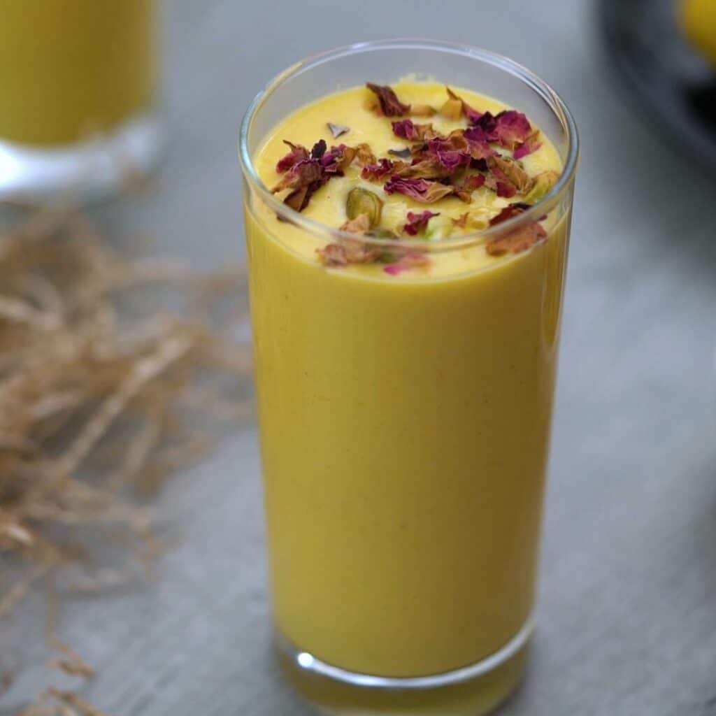 Mango Lassi served in a serving glass topped with pistachio and rose petals.