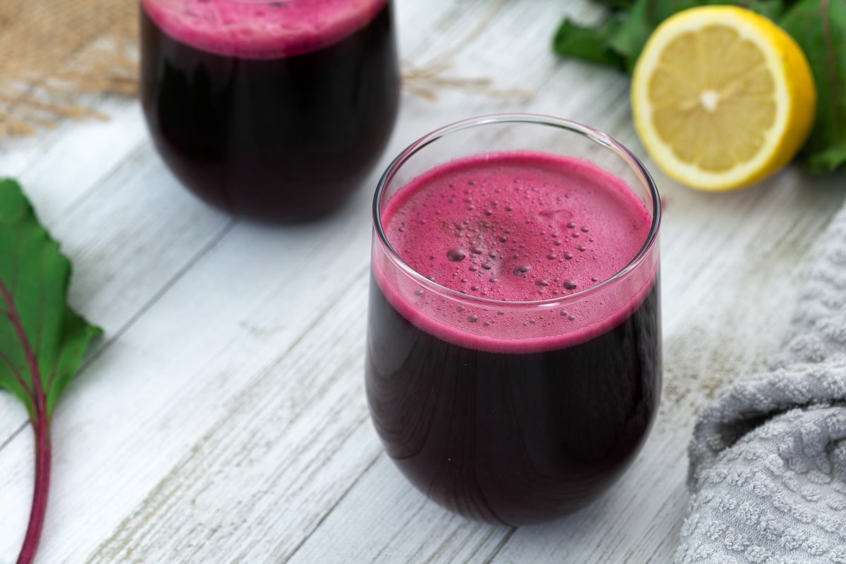 Beetroot juice in glass placed on a whiteboard with lemon and beet leaf alongside.