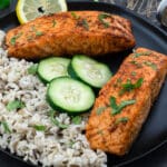 Air fryer salmon fillets in a black plate with rice and cut cucumber pieces.
