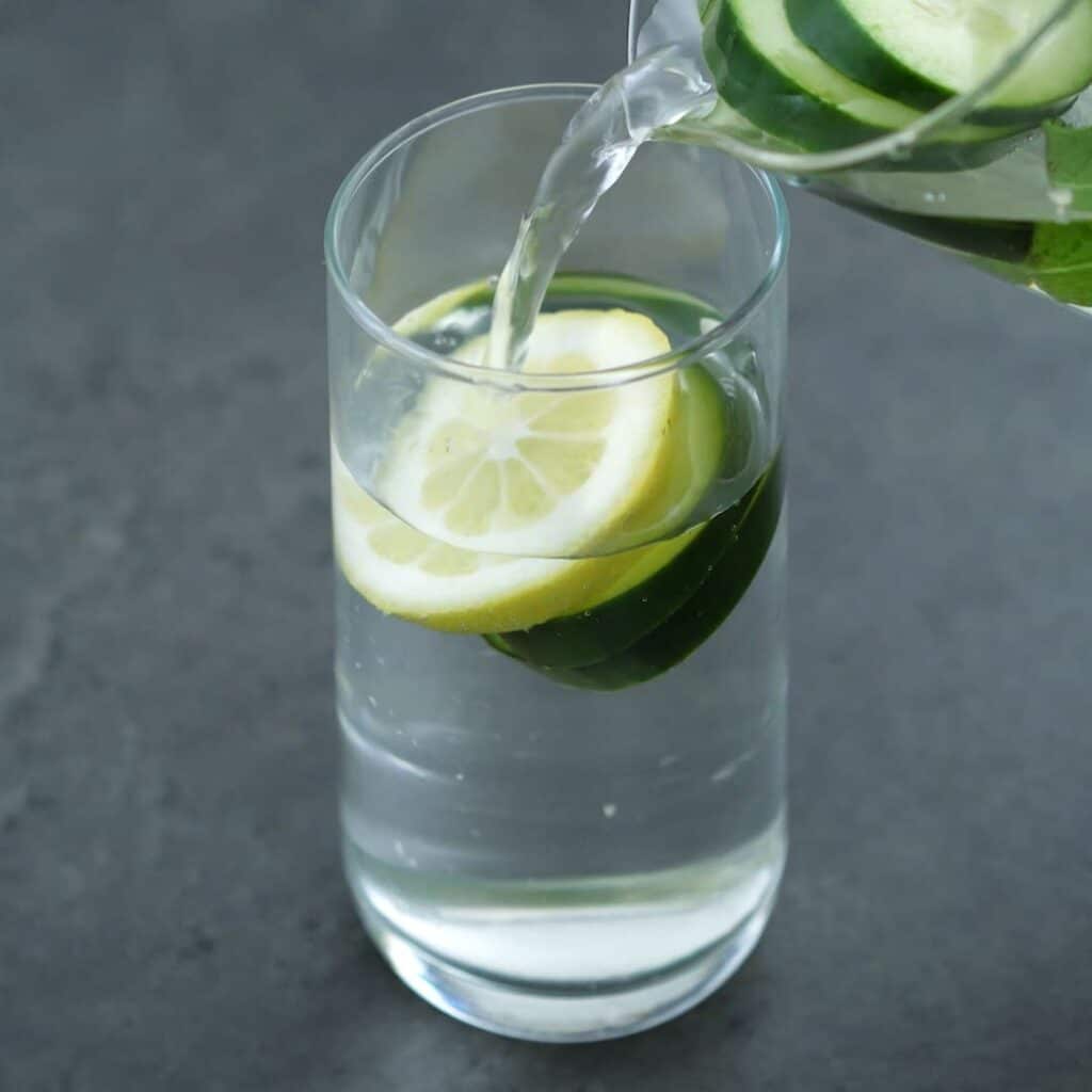 Pouring refrigerated Cucumber Water into a serving glass with cucumber and lemon slices.