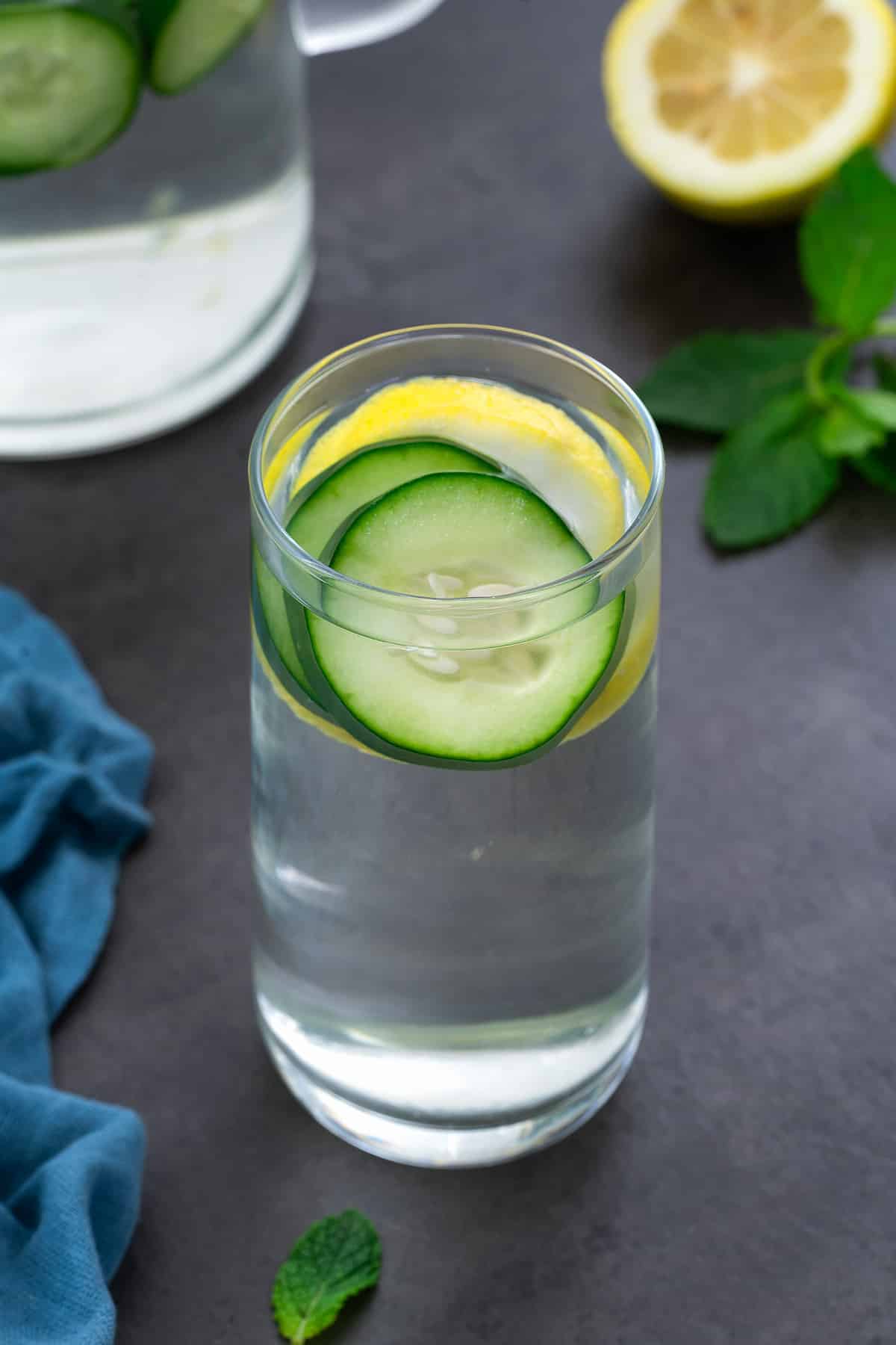 Cucumber Water in a glass placed on a grey table with lemon and mint leaves around.