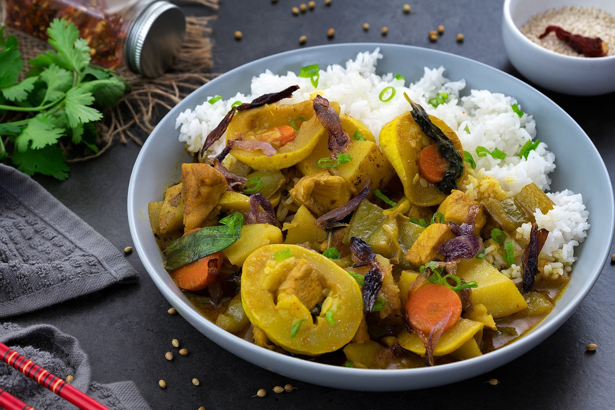 Thai Yellow Chicken Curry in a white bowl with rice placed on a grey table with few ingredients alongside