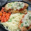 Baked Chicken Parmesan in a black plate with tomato and pasta.