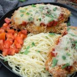 Baked Chicken Parmesan in a black plate with tomato and pasta.