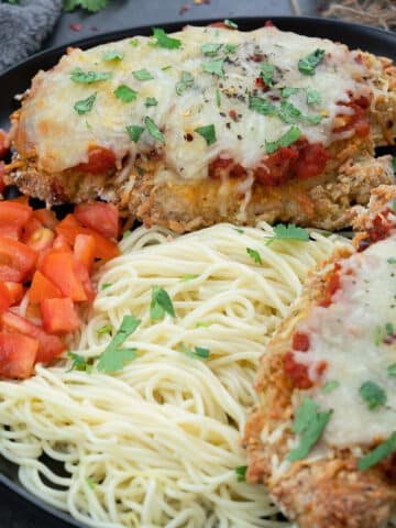 Baked Chicken Parmesan in a black plate with tomato and pasta.