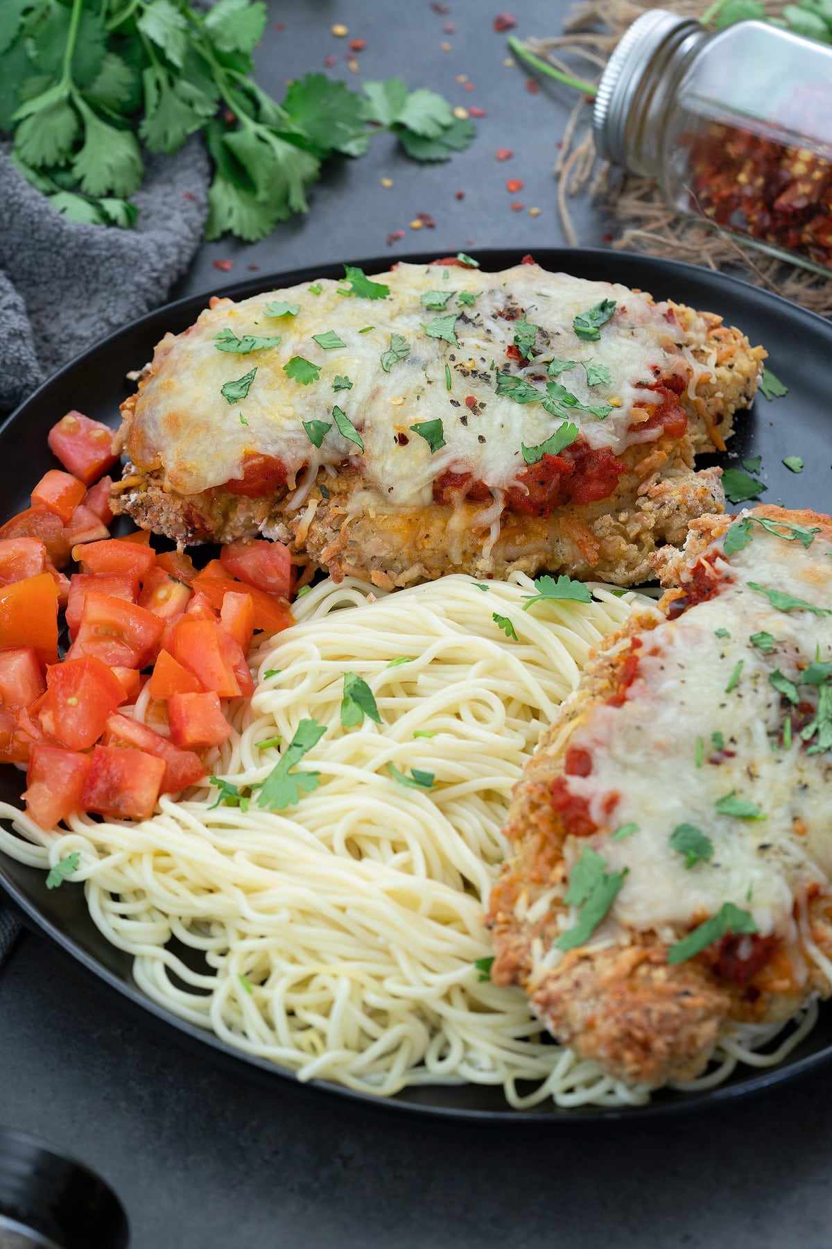 Baked Chicken Parmesan in a black plate with tomato and pasta.