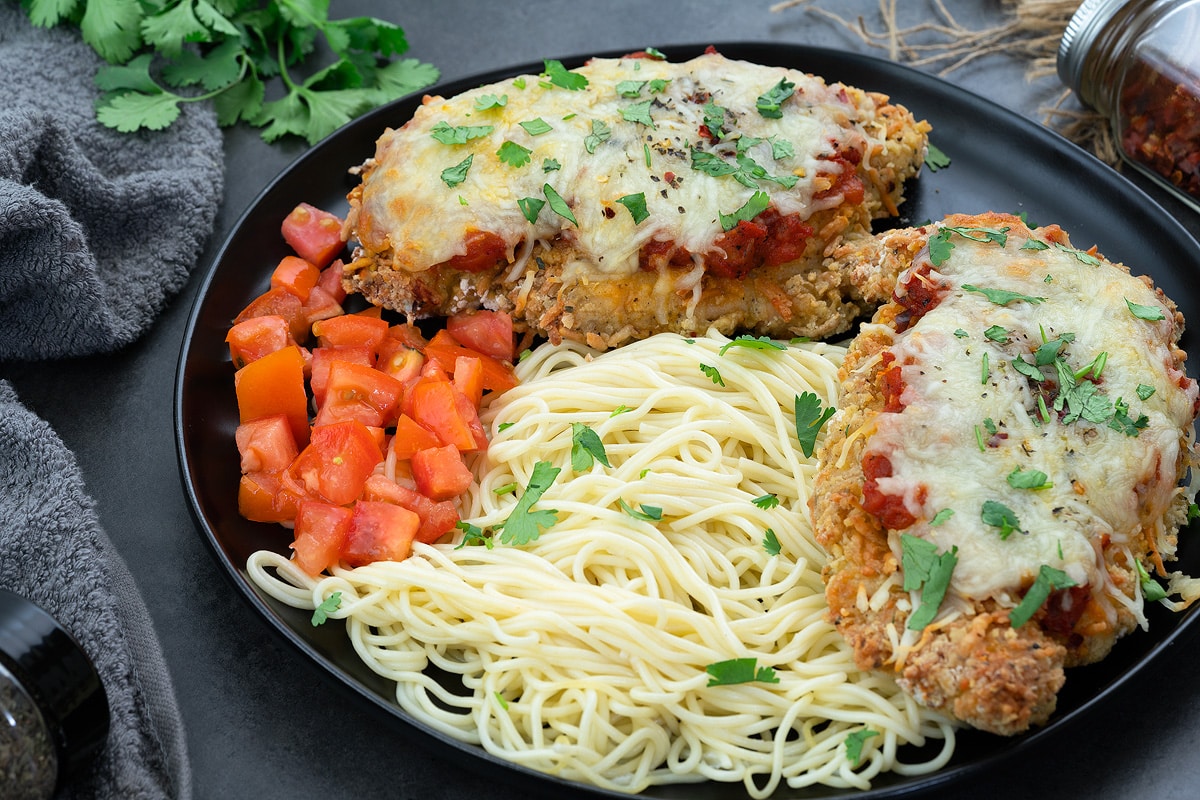 Baked Chicken Parmesan in a black plate with tomato and pasta.