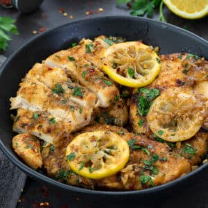 Lemon Chicken in a black bowl with lemon slices and few ingredients placed around.