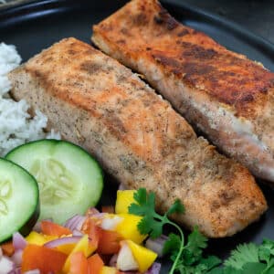 Pan fried salmon in a black plate with rice, cucumber and cut fruits.