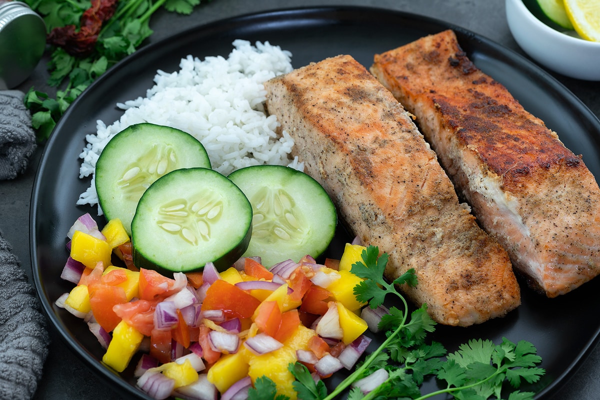 Pan fried salmon fillets in a black plate with rice, cucumber and cut fruits.