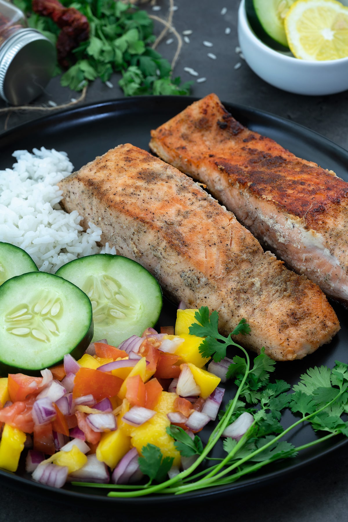 Pan fried salmon fillets in a black plate with rice, cucumber and cut fruits.