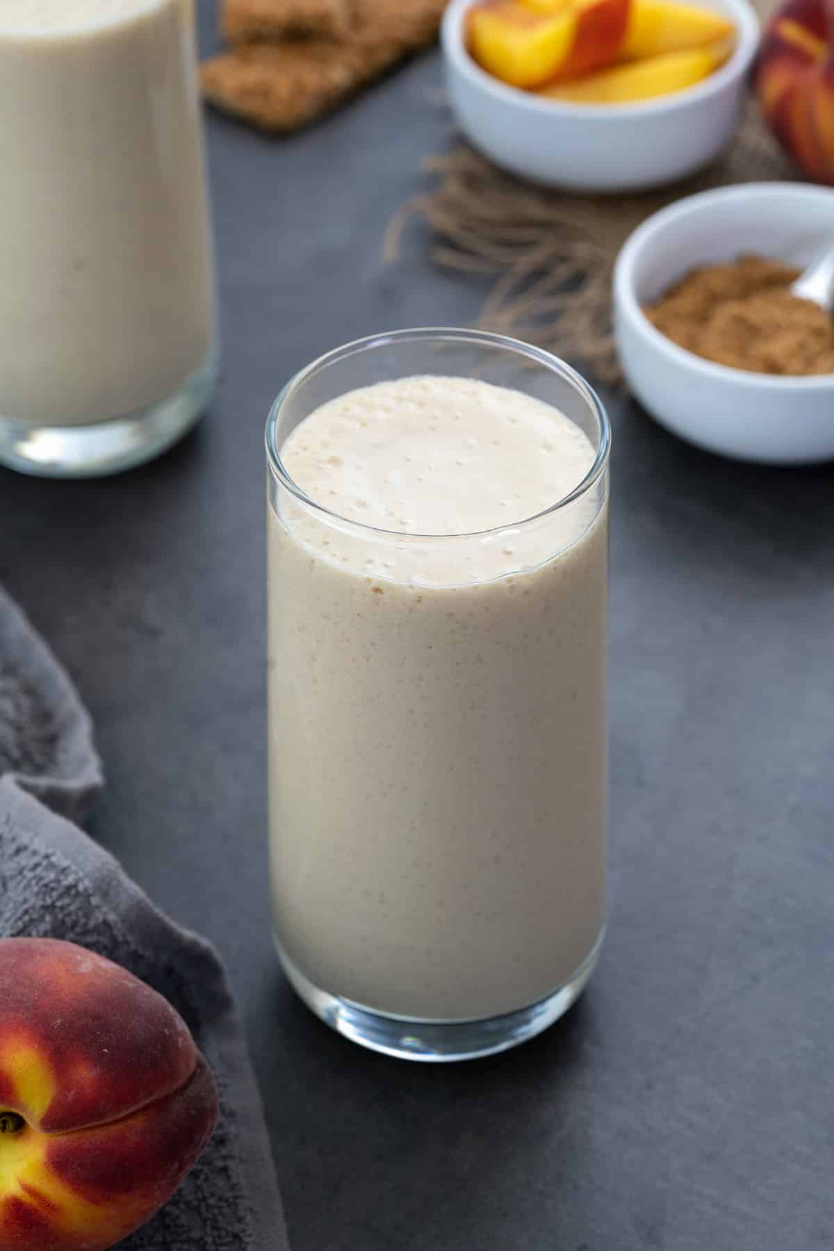 Peach smoothie in a glass with whole and cut peach with sugar around.