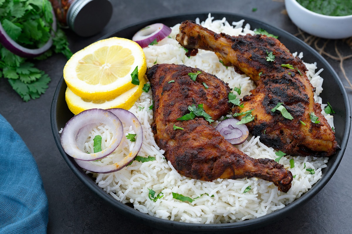Tandoori Chicken in a black bowl with rice, lemon and onion slices.