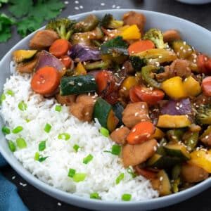 Chicken Stir Fry in a white bowl with rice with few ingredients around.