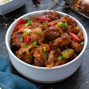 Chilli Chicken in a white bowl on a grey table with few ingredients around.