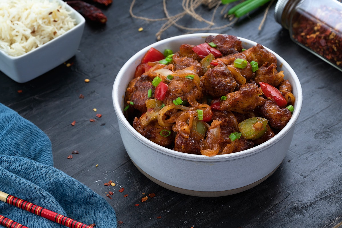 Chilli Chicken in a white bowl on a grey table with few ingredients around.