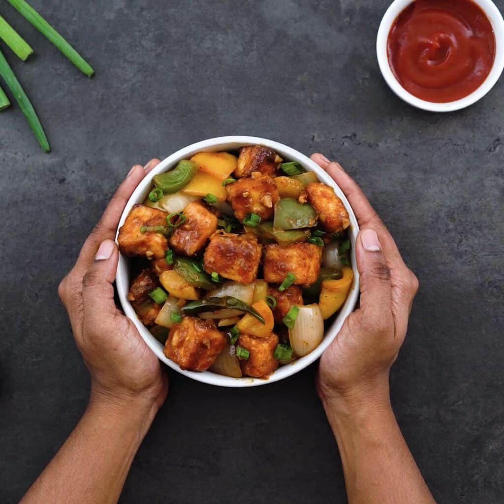 Serving Chilli Paneer in a white bowl with ketchup and spring onions alongside.