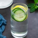 Cucumber water in a glass placed on a grey table with few ingredients around.