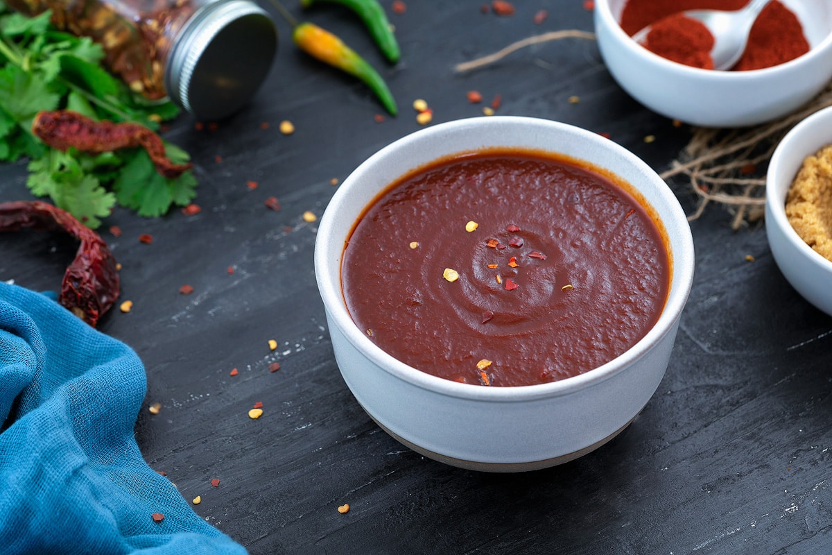Spicy homemade chili sauce in a white bowl on a grey table with few ingredients around.