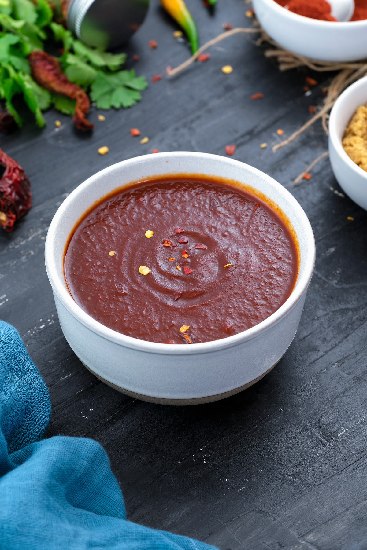 Spicy homemade chili sauce in a white bowl on a grey table with few ingredients around.