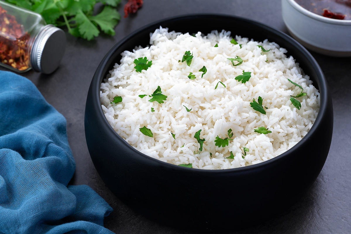 Perfectly cooked fluffy white rice in a black bowl on a grey table, with few ingredients around.