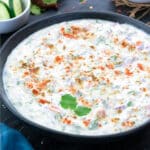 Raita in a black bowl placed on a grey table with few ingredients around.