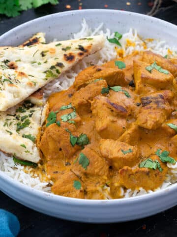 A white bowl filled with butter chicken curry, along with rice and naan.