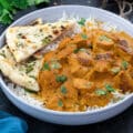 Butter chicken curry served in a bowl with rice and naan, and a few ingredients on the side.