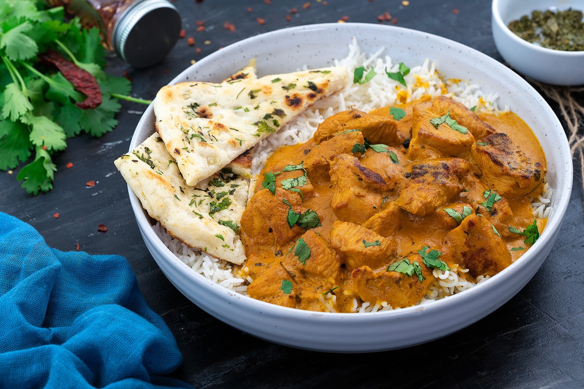 Butter chicken curry served in a bowl with rice and naan, and a few ingredients on the side.