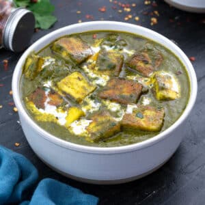 A white bowl containing saag paneer, with few ingredients scattered around it.