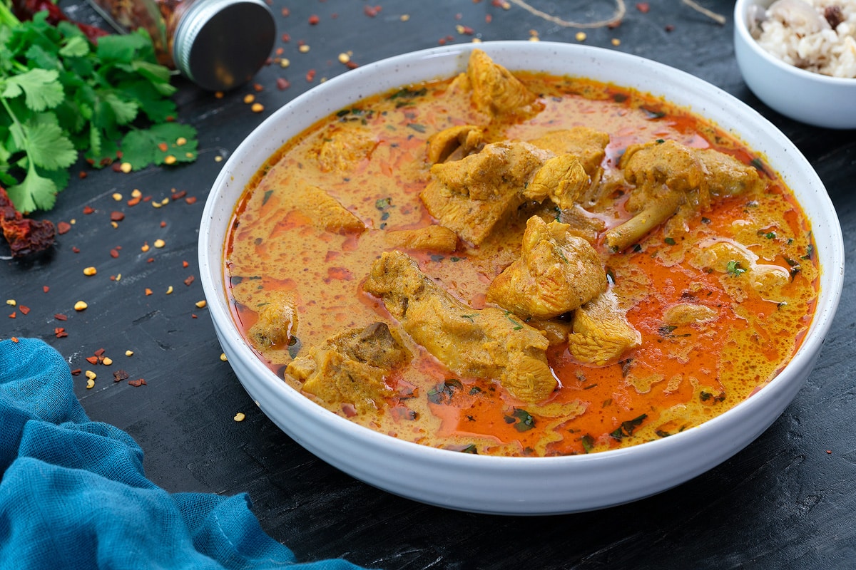 A bowl of chicken korma on a grey table with few ingredients scattered around.