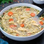 Chicken noodle soup in a white bowl with few ingredients scattered around.