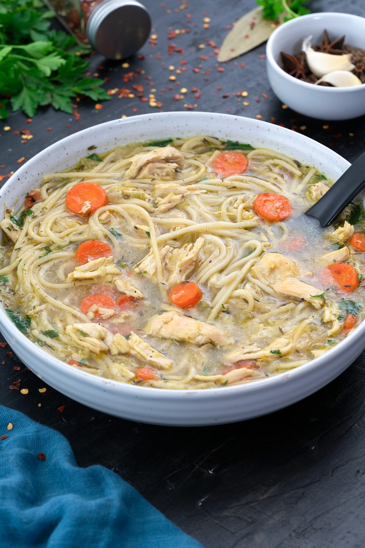 Chicken noodle soup in a white bowl with few ingredients scattered around.