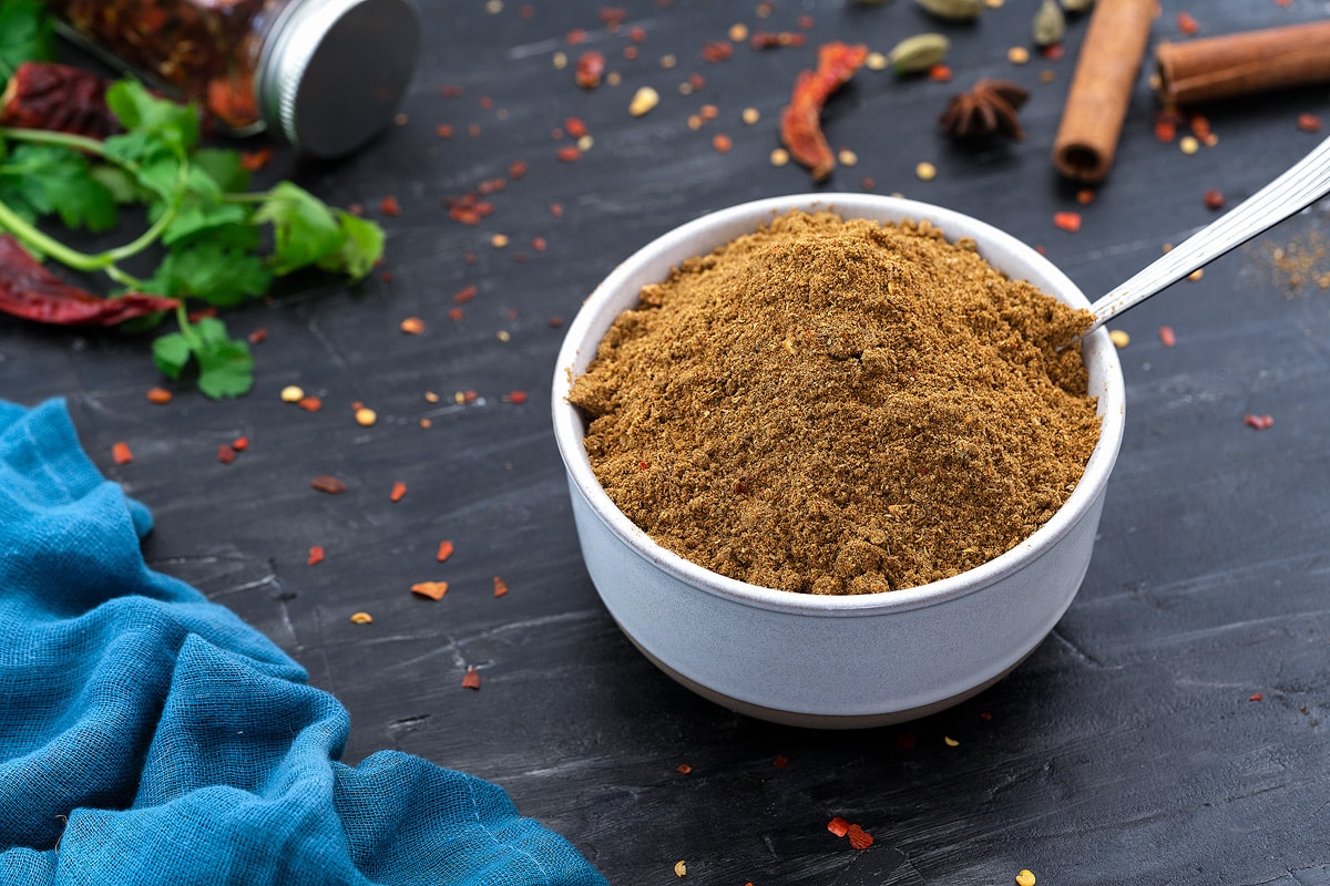 A white cup of Garam Masala spice mix, with various ingredients such as whole spices scattered around it.