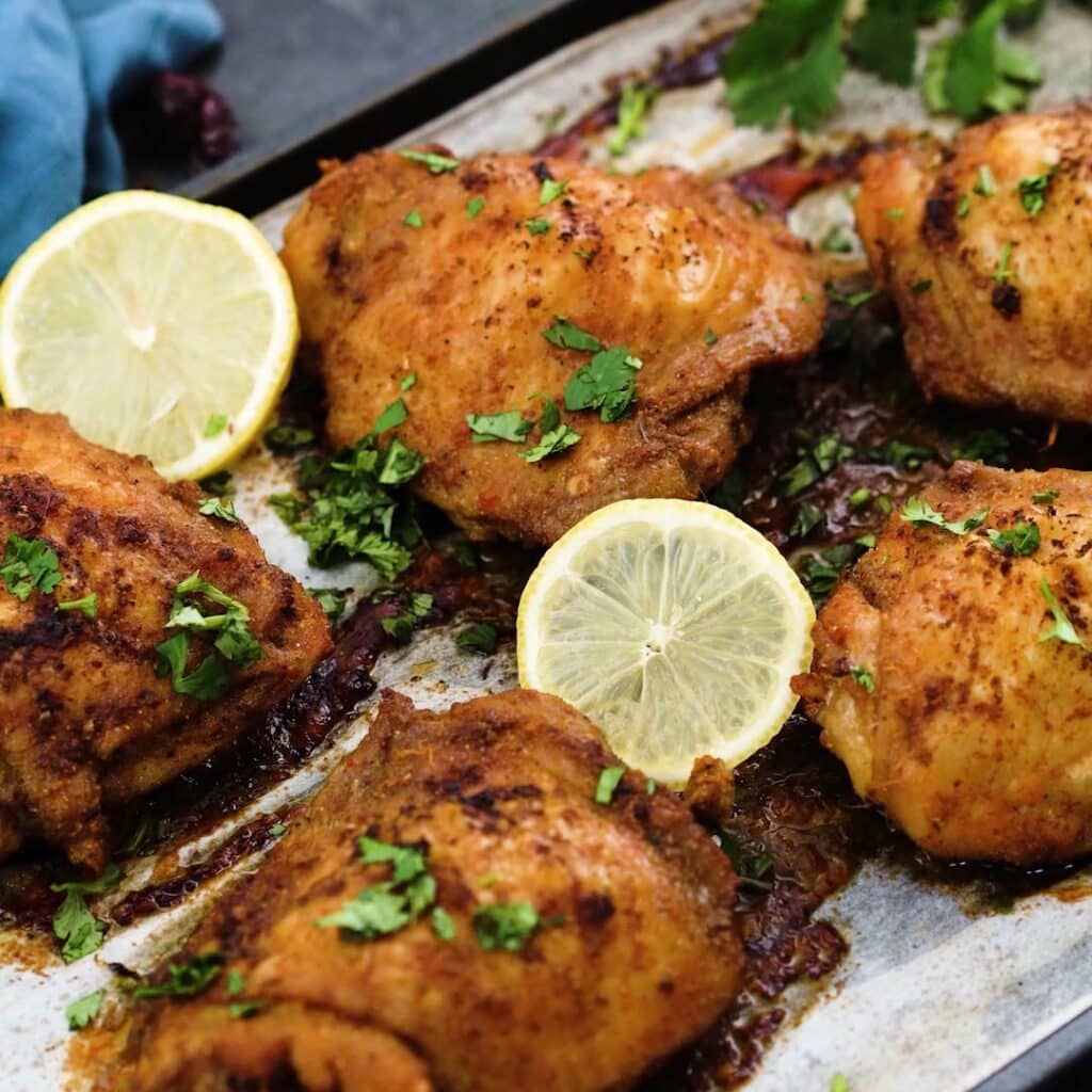 Oven Baked Chicken thighs on a baking tray garnished with coriander leaves and lemon slice.