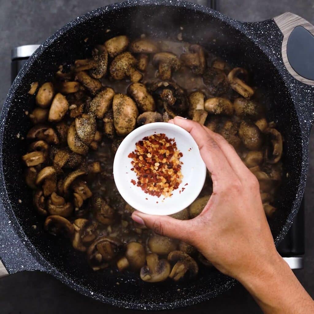 Adding chili flakes to the pan with mushroom.