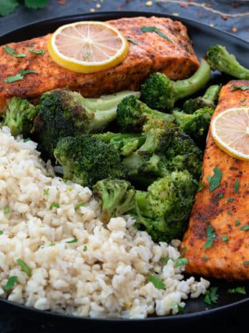 Baked Salmon fillets in a black plate with brown rice and broccoli.
