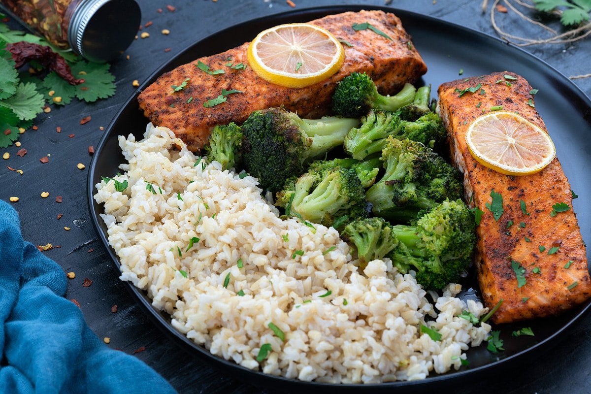 Baked Salmon fillets in a black plate with brown rice and brocolli.