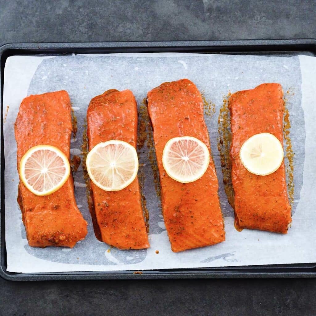 A baking tray with marinated salmon that is topped with lemon slices.