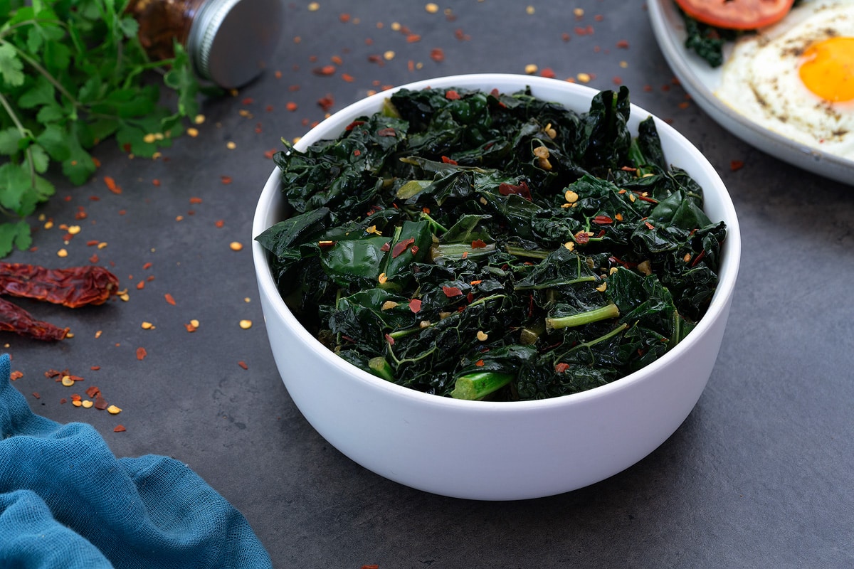 Sauteed Kale in white bowl with few ingredients scattered around.