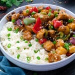 Sweet and Sour Chicken served in a white bowl along with rice with few ingredients scattered around.