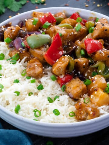 Sweet and Sour Chicken served in a white bowl along with rice with few ingredients scattered around.
