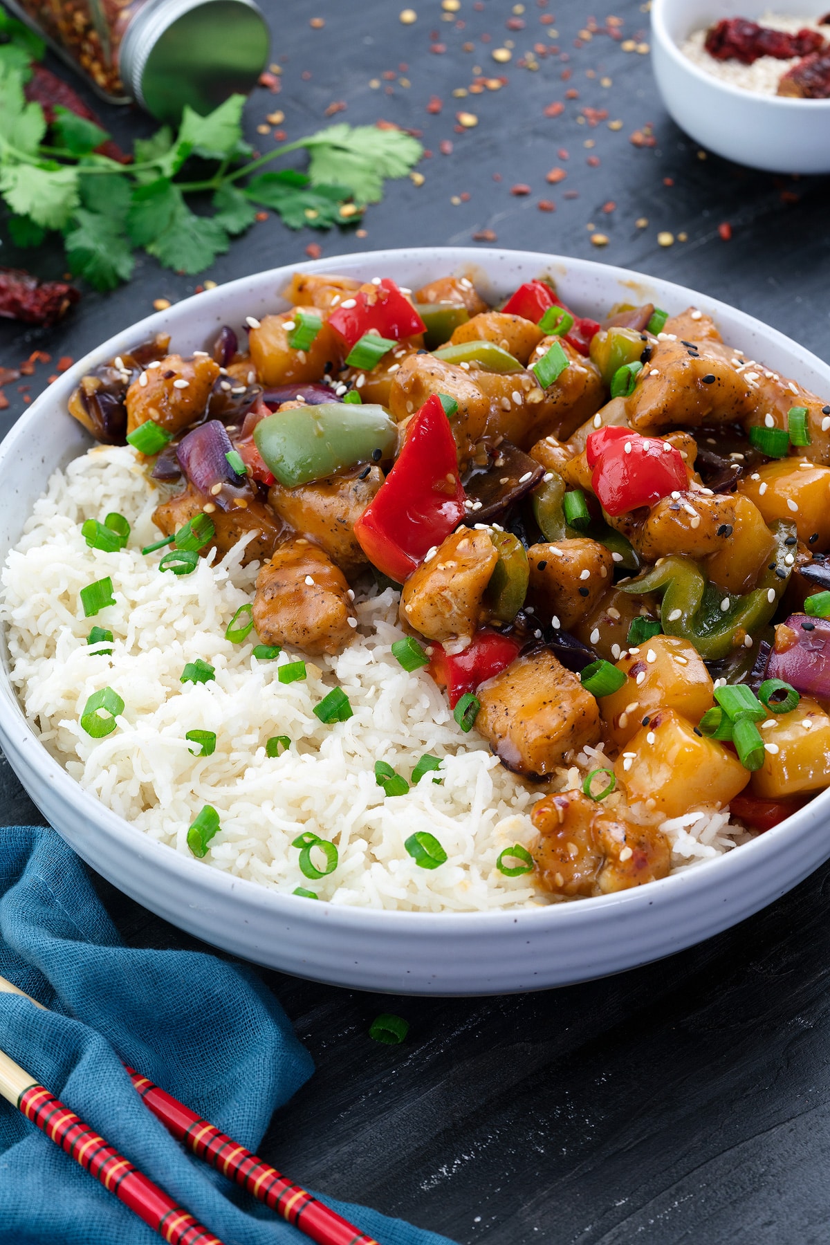 Sweet and Sour Chicken served in a white bowl along with rice with few ingredients scatterred around.