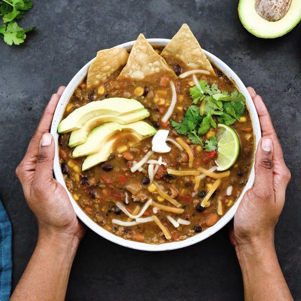 Serving Turkey Chili topped with avocado, tortilla chips, cheese blend, sour cream and lime wedges in a bowl.