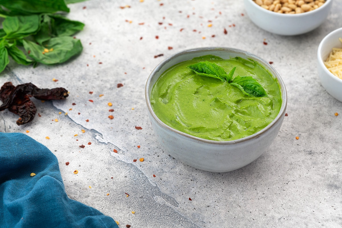 Basil Pesto Sauce in a white bowl with few ingredients scattered around.