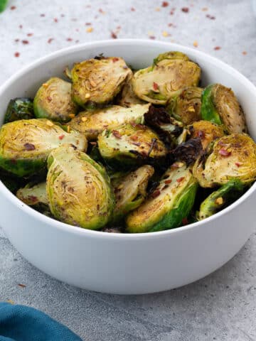 Air fryer brussels sprouts served in a while bowl on a white table with few ingredients scattered around.