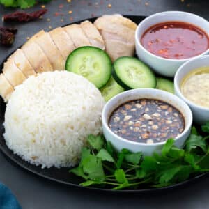 Hainanese Chicken Rice served with different dipping sauces in a black plate, along with few ingredients scattered around the plate.