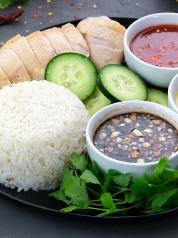 Hainanese Chicken Rice served with different dipping sauces in a black plate, along with few ingredients scattered around the plate.