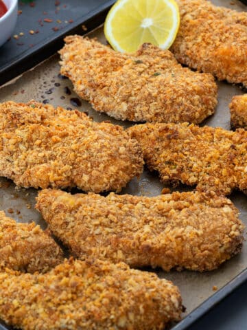 Oven Fried Chicken Tenders in a baking tray with hot sauce and lemon slice along side.