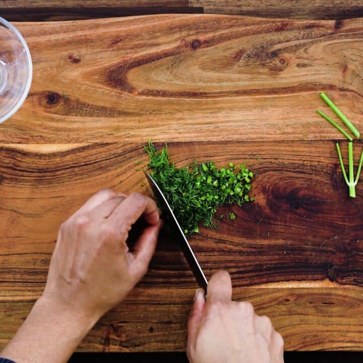 Chopping the dill leaves on wooden board using knife.